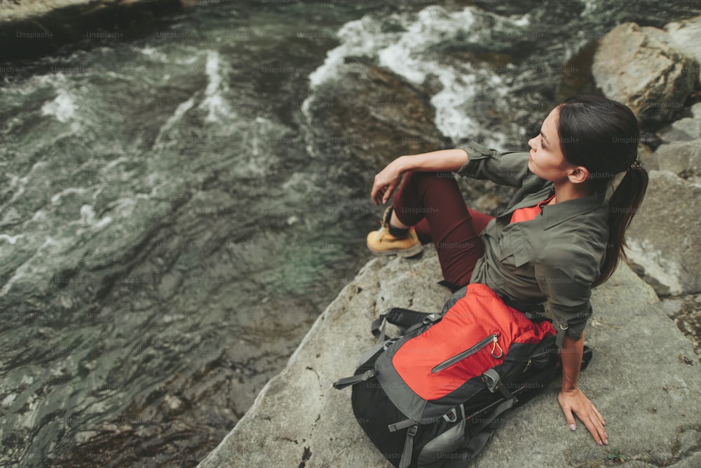 Delighted young woman sitting on the bank of stone river while having an active weekend